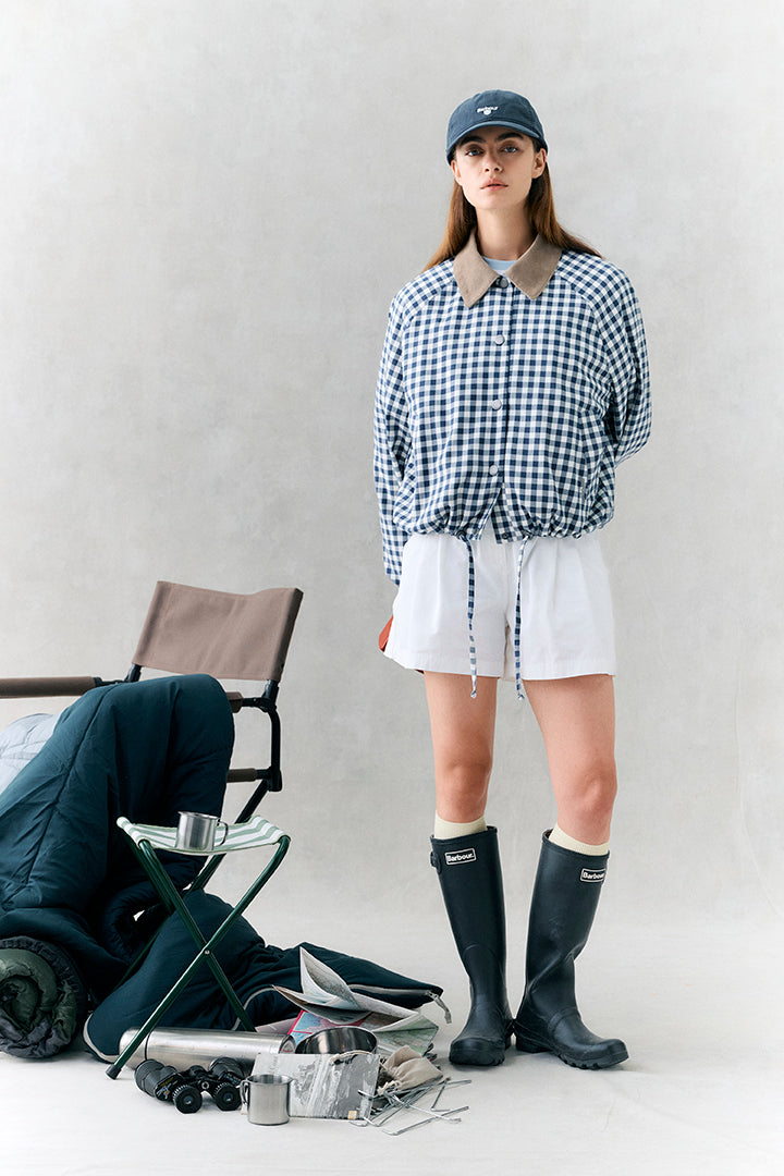 Person wearing a checkered shirt, white shorts, and black rain boots with a cap, standing next to outdoor gear on a light background.