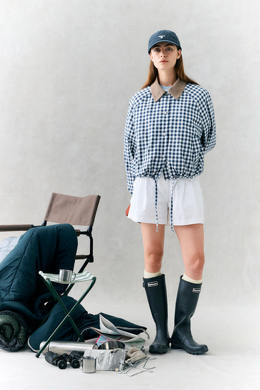 Person wearing a checkered shirt, white shorts, and black rain boots with a cap, standing next to outdoor gear on a light background.
