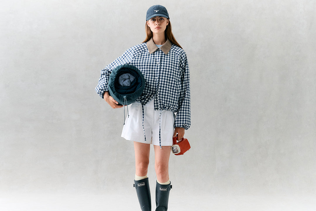 Person wearing a checkered shirt, white shorts, and rain boots holding a rolled-up mat and bag against a plain background