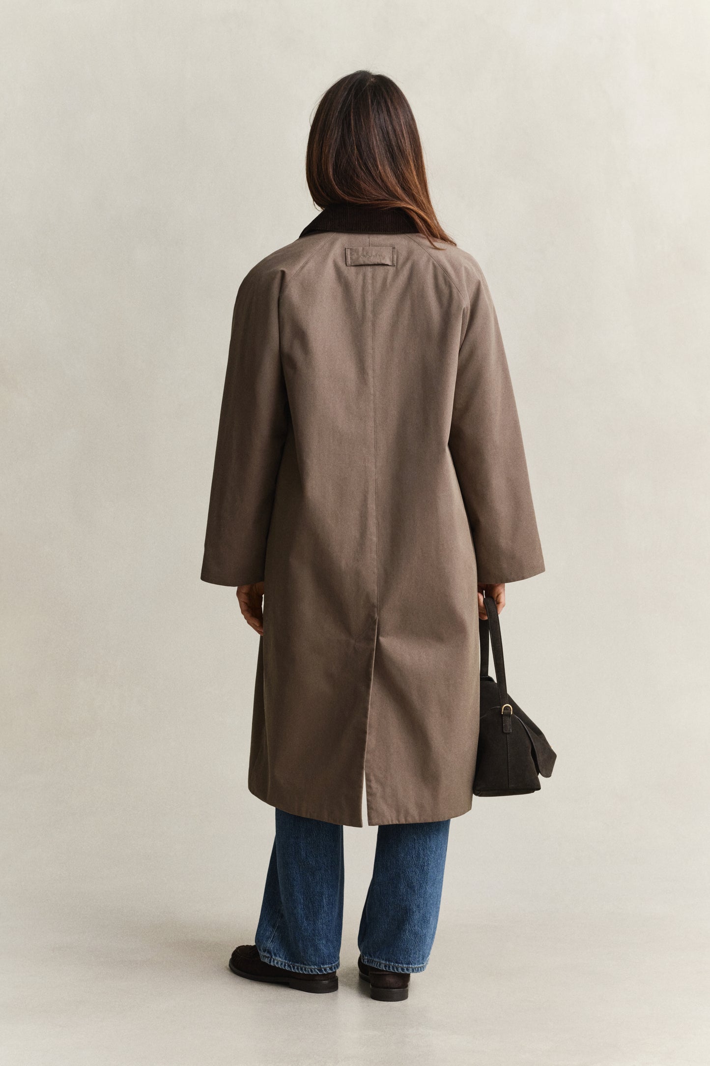 Person wearing a brown coat and blue jeans, standing against a plain background
