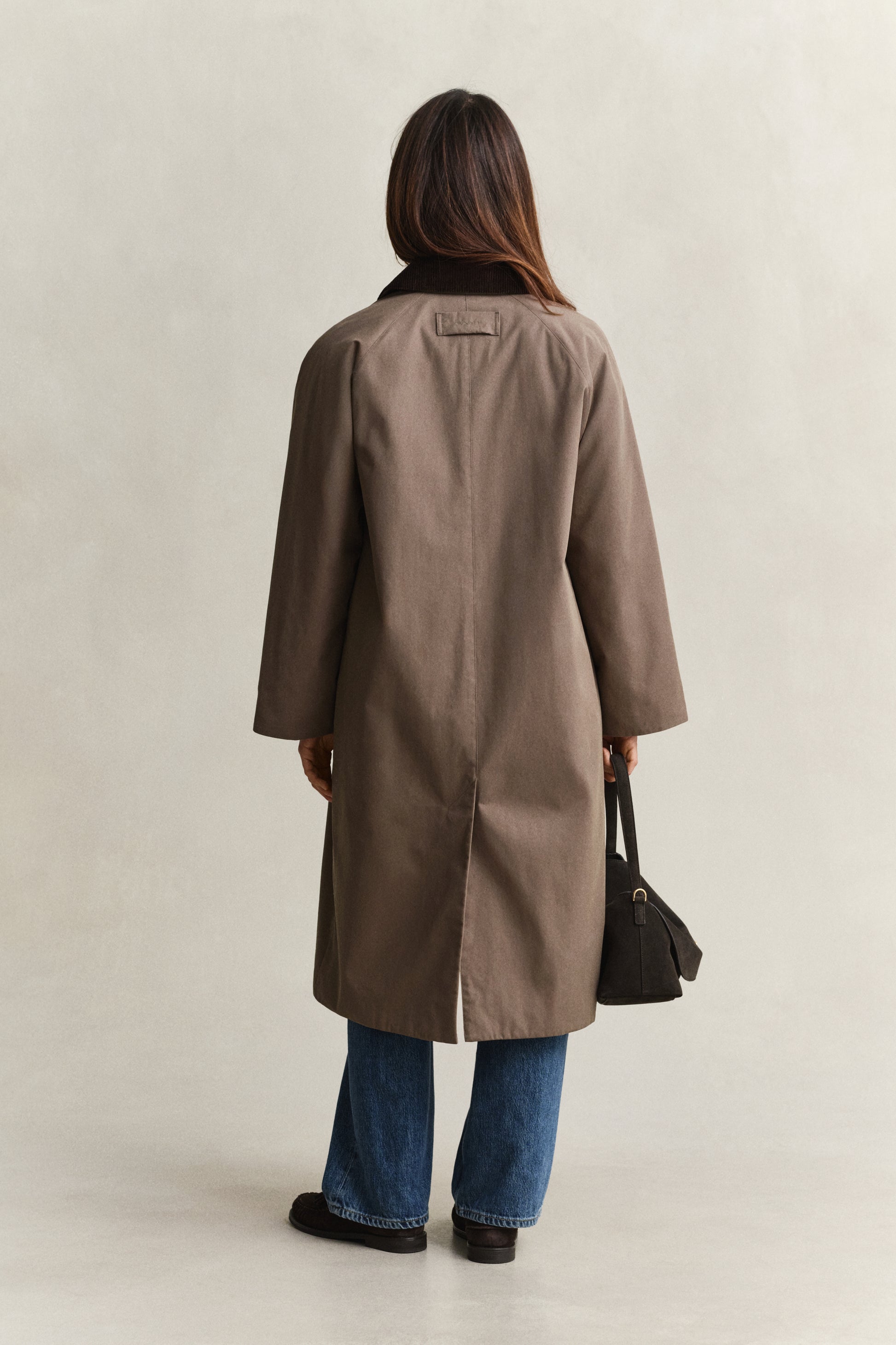 Person wearing a brown coat and blue jeans, standing against a plain background
