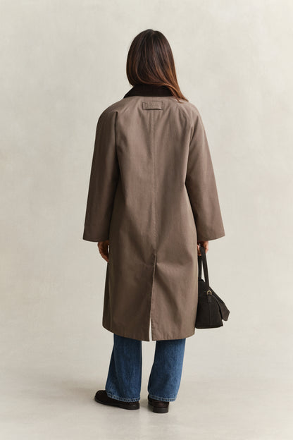 Person wearing a brown coat and blue jeans, standing against a plain background