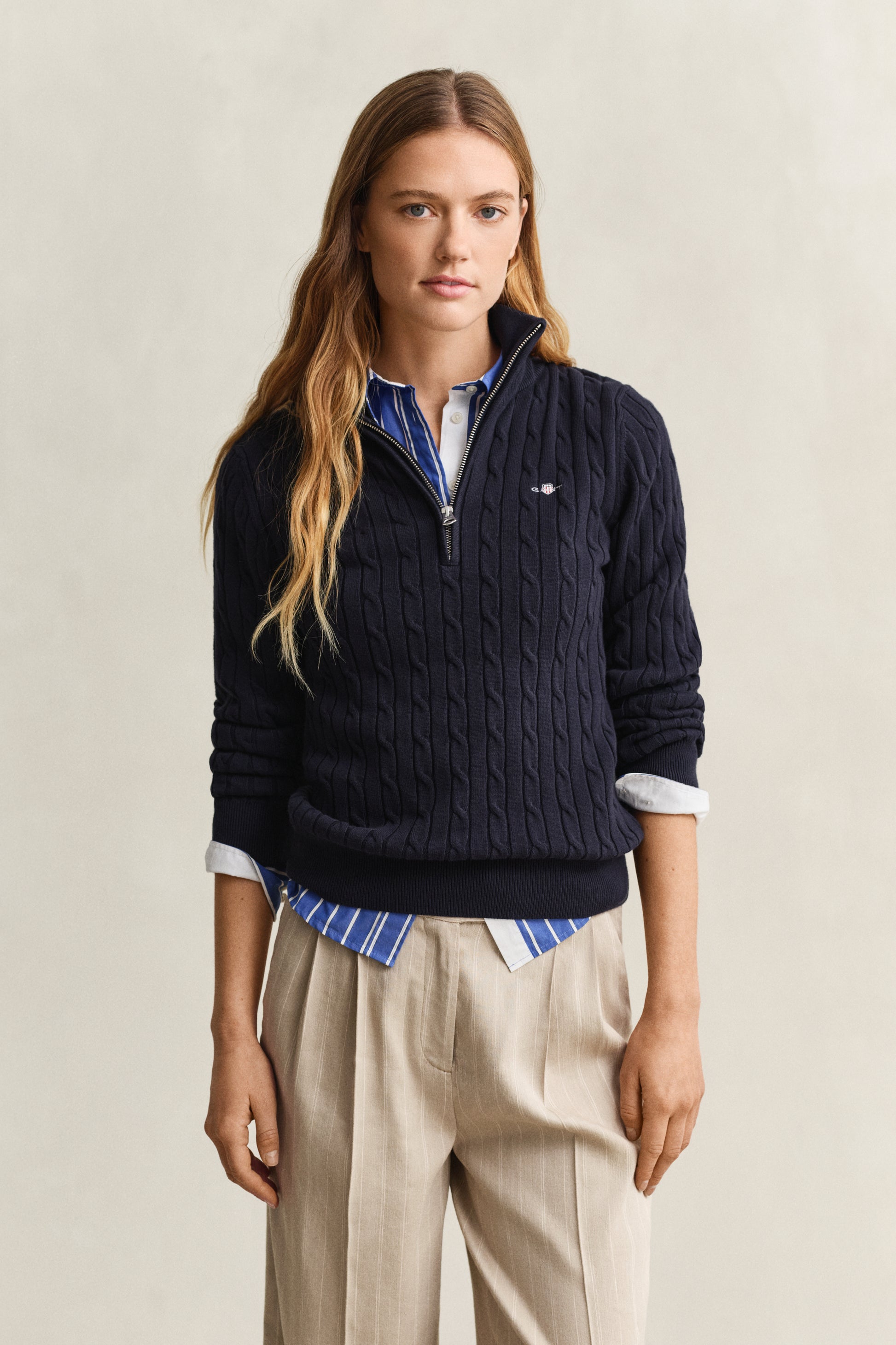 Woman wearing a navy half-zip sweater over a striped shirt with beige pants on a beige background