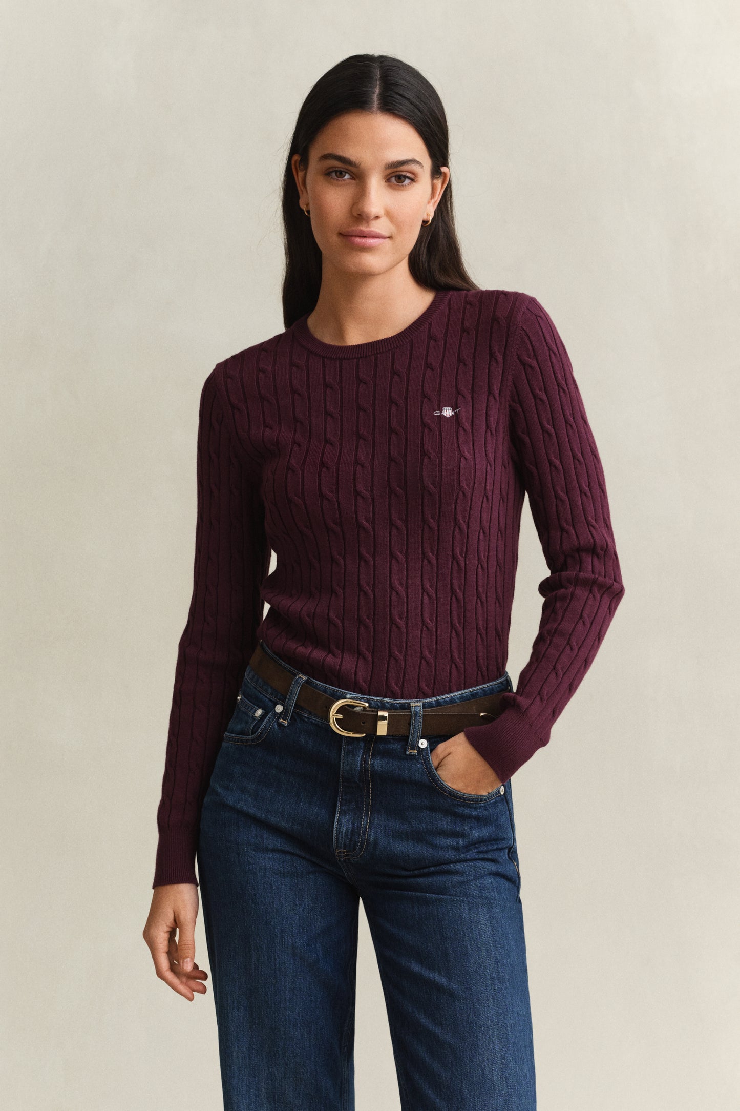 Woman wearing a maroon sweater with a logo and blue jeans on a beige background
