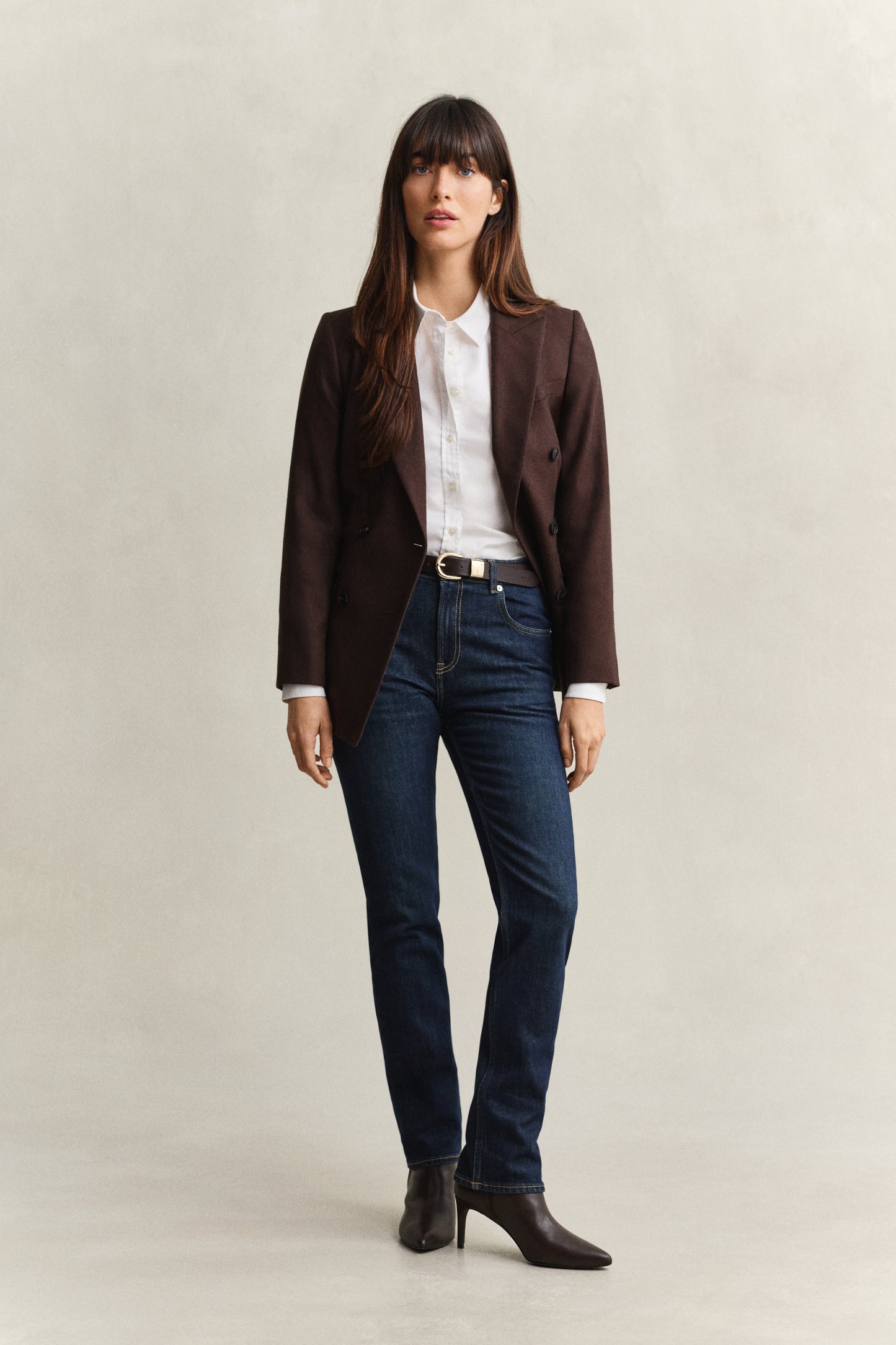 Woman wearing a brown blazer, white shirt, and blue jeans on a beige background