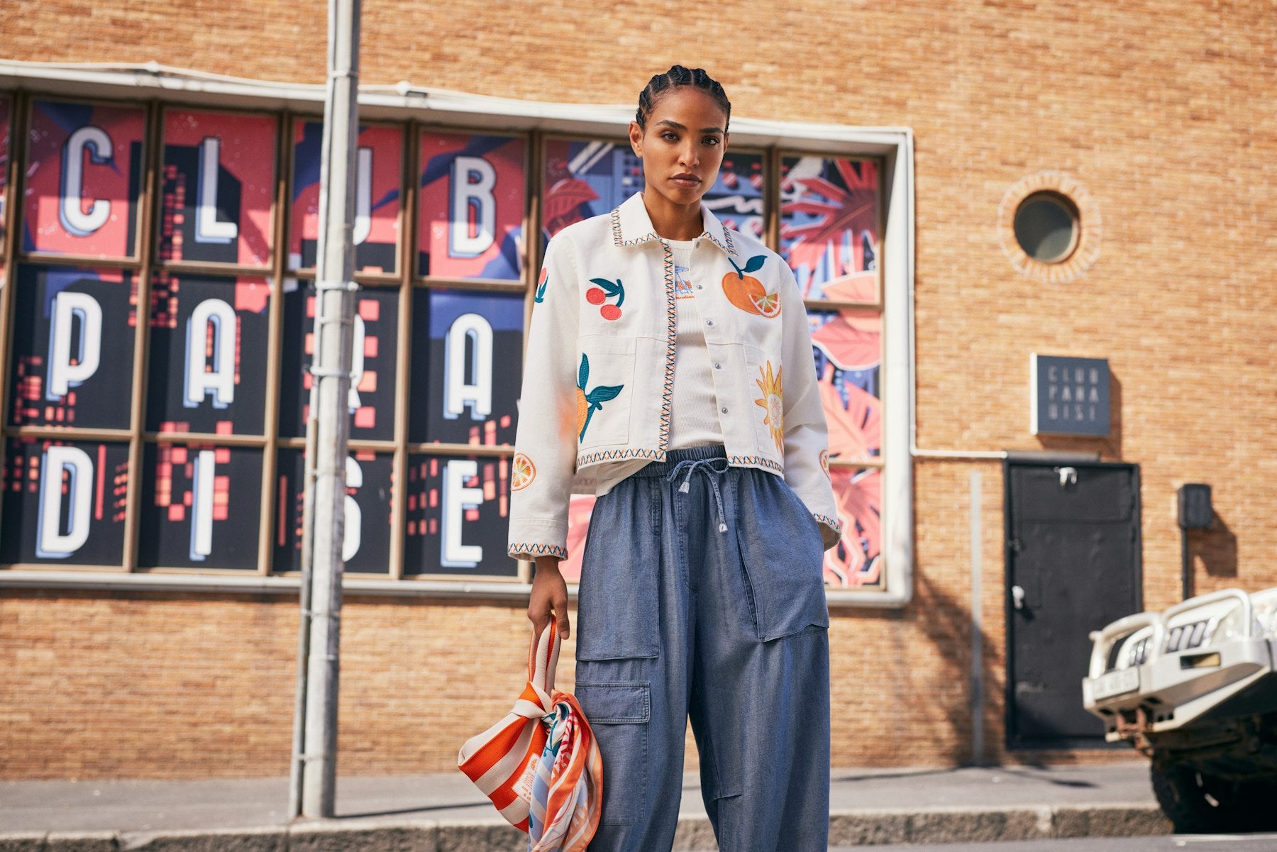 Woman in an embroidered jacket and blue pants standing in front of a brick building with colorful window display.