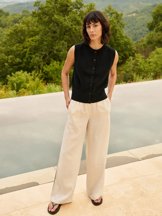 Woman wearing a black sleeveless top and white pants standing outdoors with greenery in the background