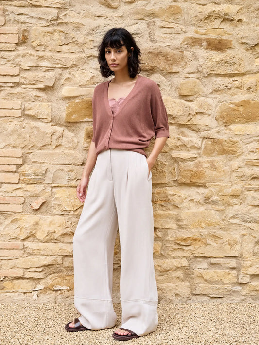 Woman wearing a pink top and white pants standing against a stone wall.