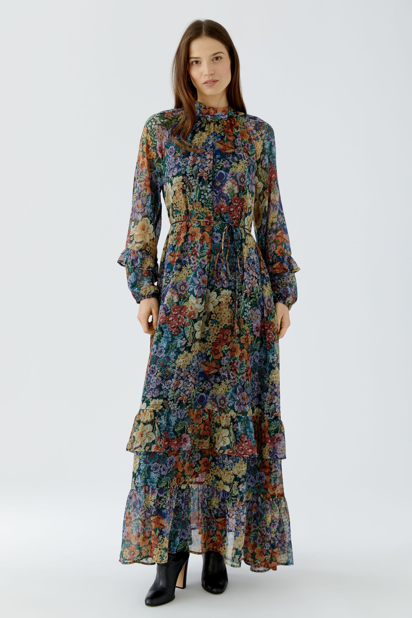A woman modeling a long-sleeved, high-neck maxi dress with a dense multicolor floral print, paired with black ankle boots, against a light gray studio background.