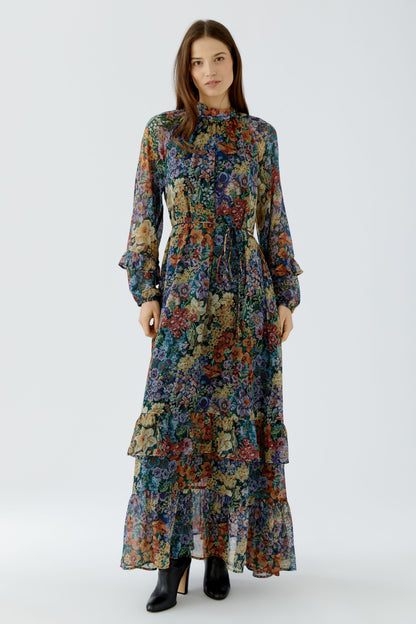 A woman modeling a long-sleeved, high-neck maxi dress with a dense multicolor floral print, paired with black ankle boots, against a light gray studio background.