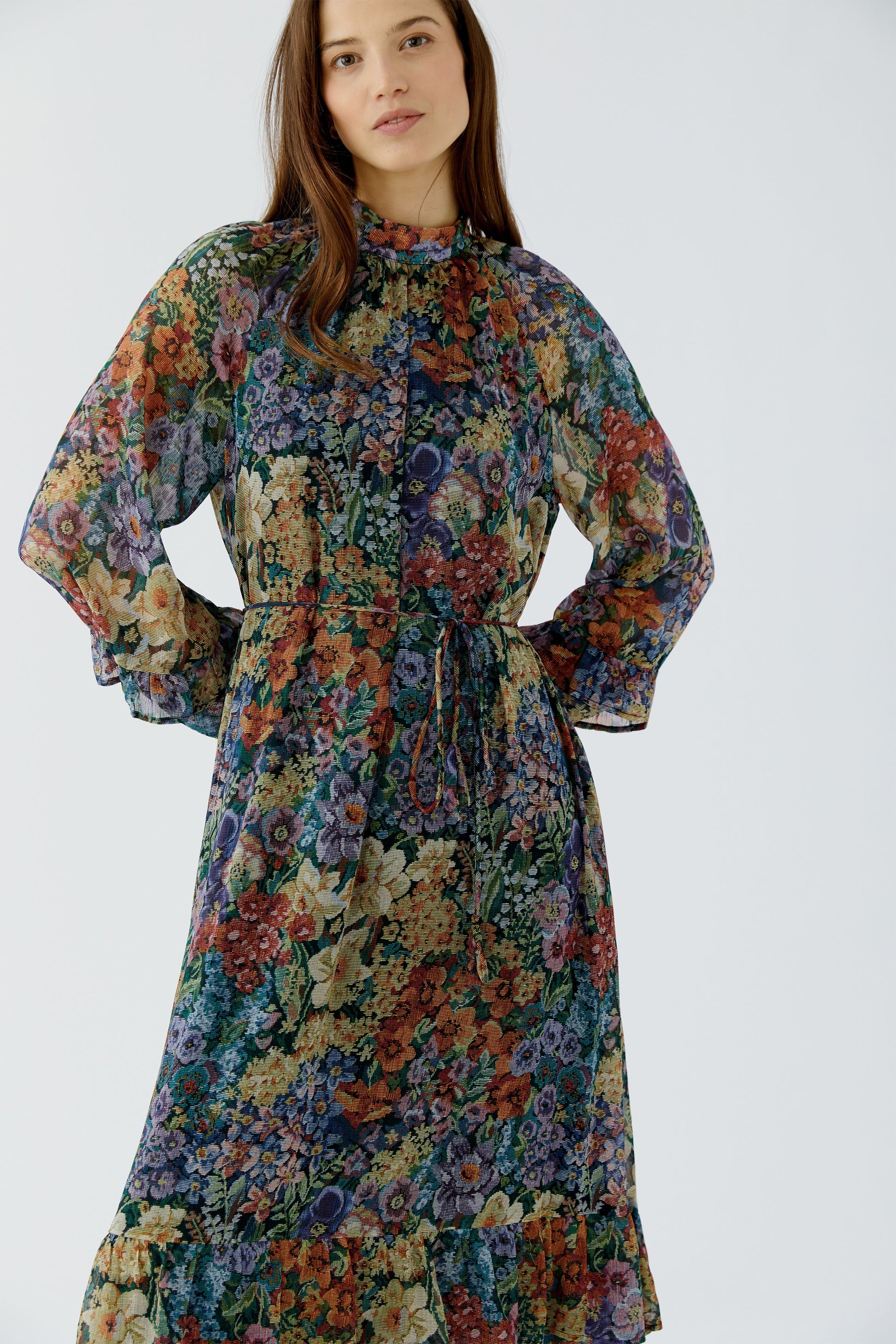 A woman wearing a long-sleeved, high-neck midi dress with a dense multicolor floral print, featuring a tie waist and ruffled hem, set against a plain light gray studio background.