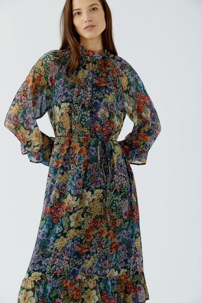 A woman wearing a long-sleeved, high-neck midi dress with a dense multicolor floral print, featuring a tie waist and ruffled hem, set against a plain light gray studio background.