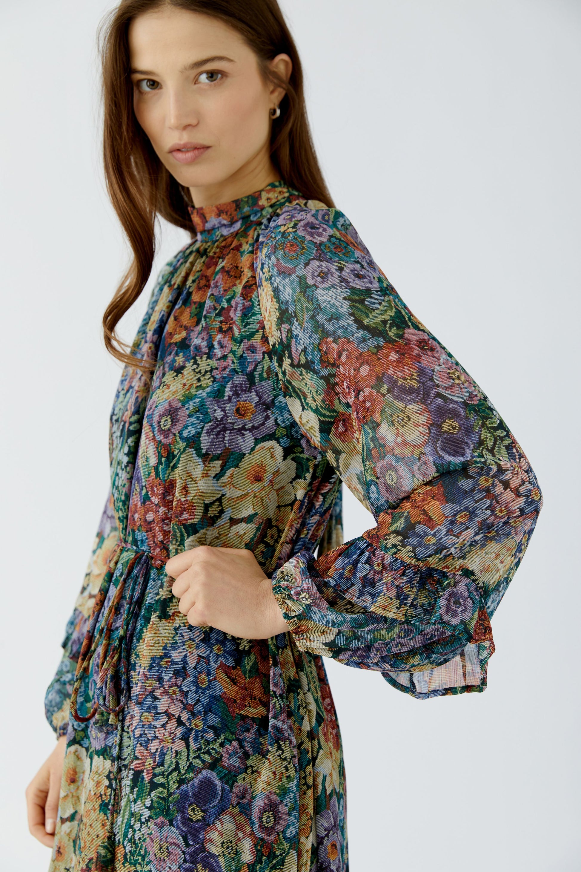 A woman wearing a long-sleeve high-neck dress with a dense multicolor floral print in blue, green, purple, red, and yellow, photographed against a light gray studio background.