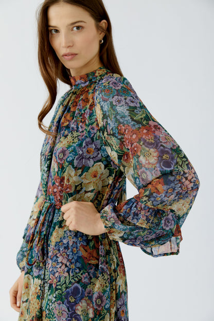 A woman wearing a long-sleeve high-neck dress with a dense multicolor floral print in blue, green, purple, red, and yellow, photographed against a light gray studio background.