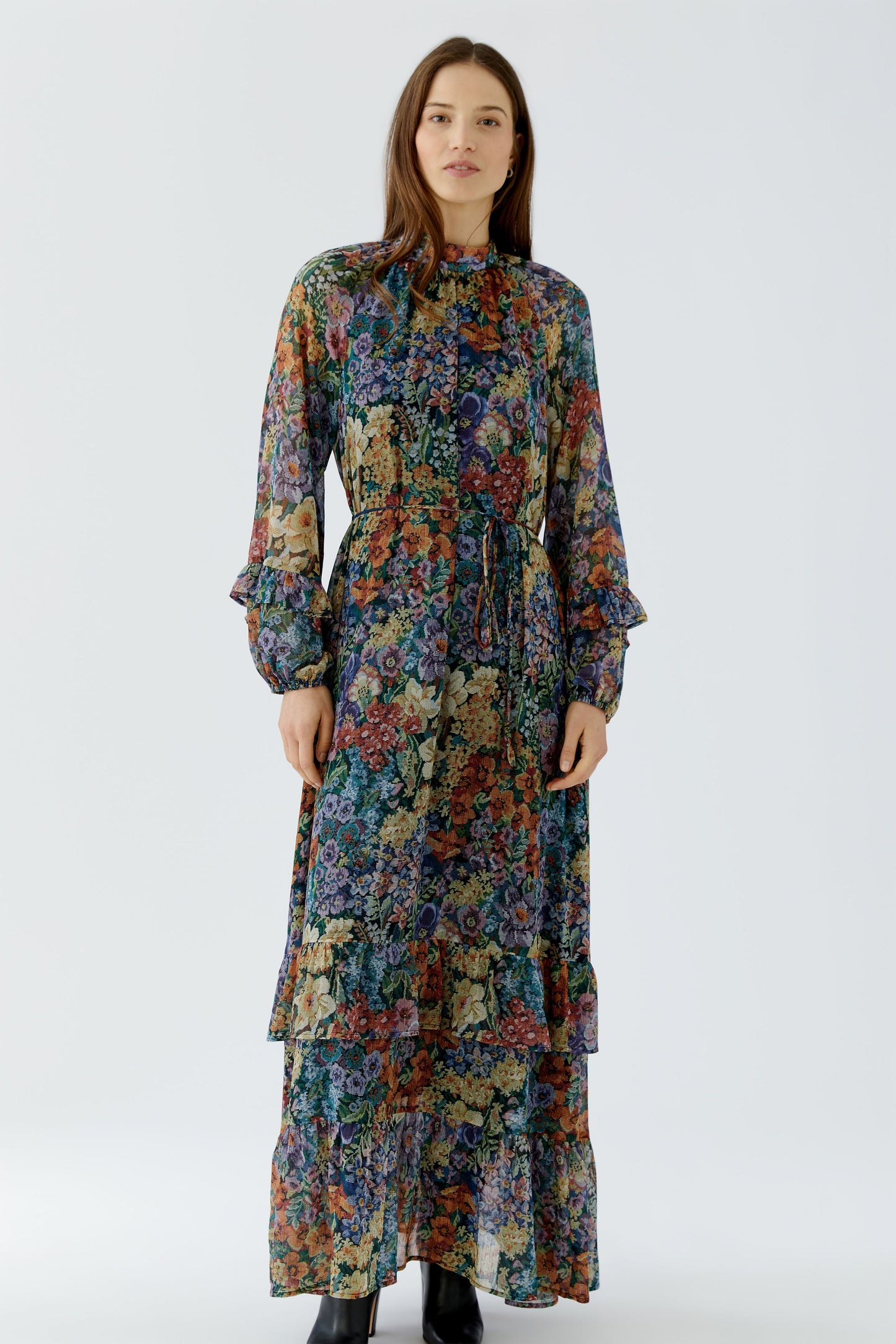 A woman modeling a long-sleeved, high-neck maxi dress with a dense multicolor floral print, standing against a plain light gray studio background.