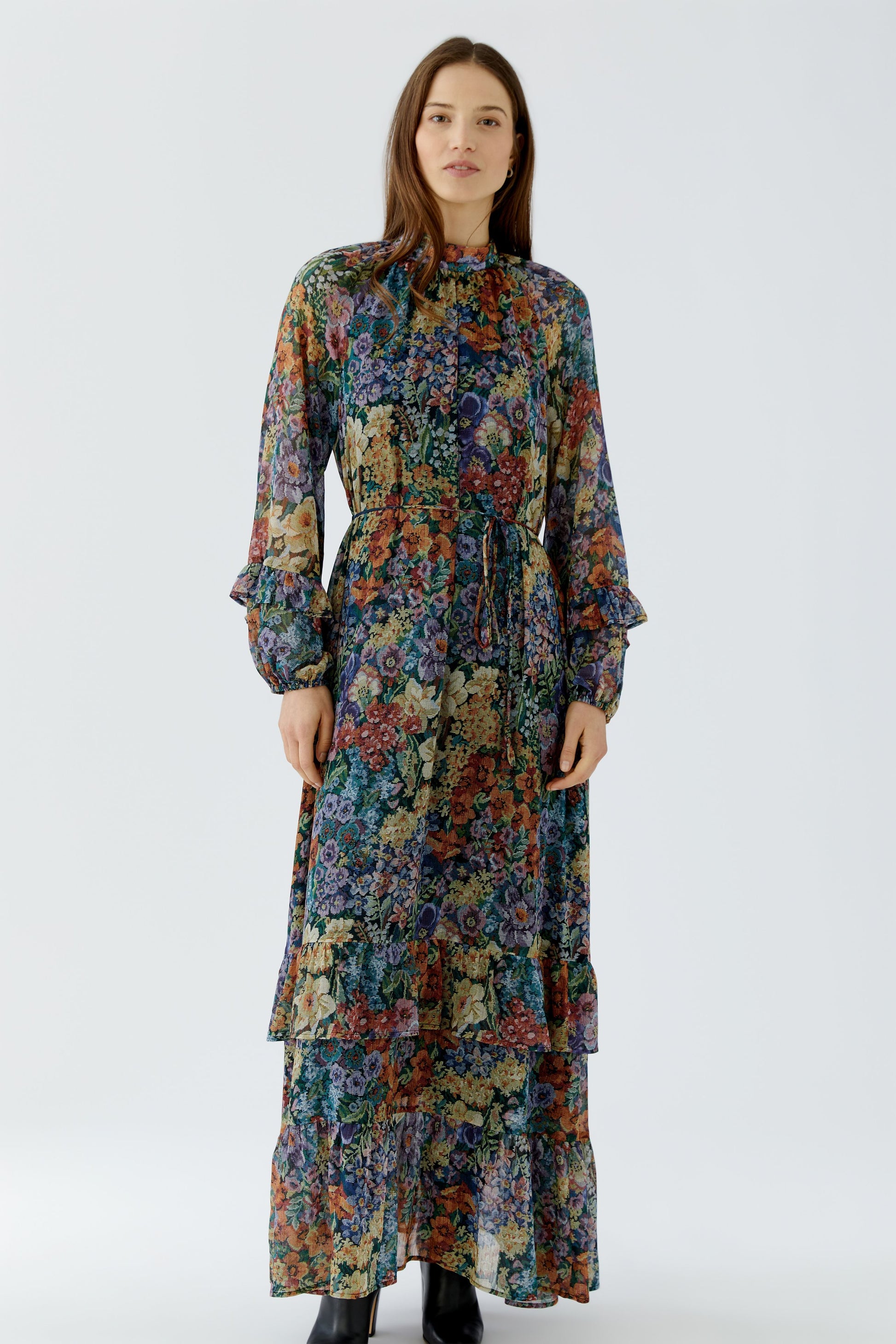 A woman modeling a long-sleeved, high-neck maxi dress with a dense multicolor floral print, standing against a plain light gray studio background.