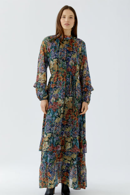 A woman modeling a long-sleeved, high-neck maxi dress with a dense multicolor floral print, standing against a plain light gray studio background.