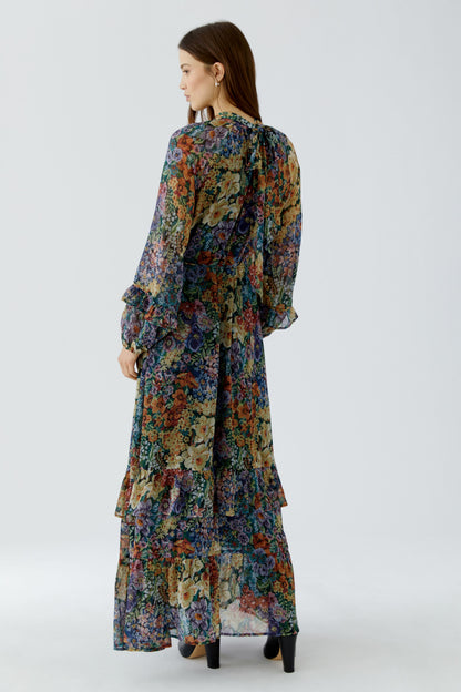 A woman modeling a long-sleeved maxi dress with a dense multicolor floral print, shown from a rear three-quarter view against a light gray studio background.