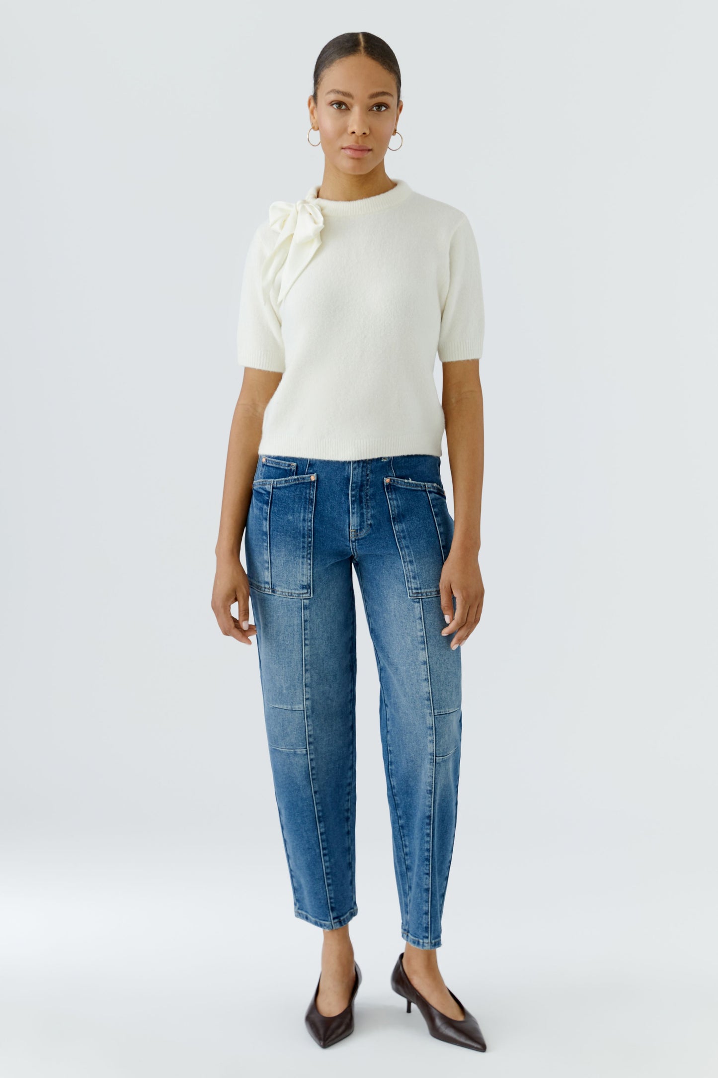 A model stands against a light gray studio background wearing a cream short-sleeve knit top with a large bow detail on the shoulder, blue cropped jeans, and dark brown pointed-toe heels.