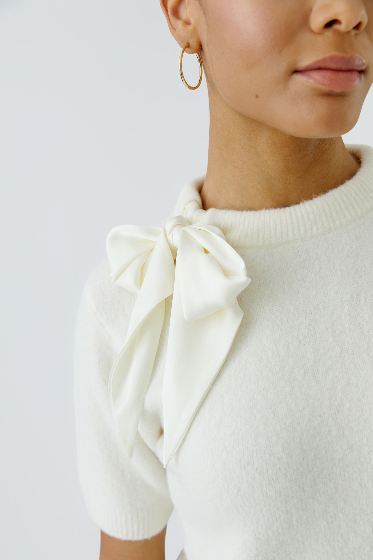 Close-up of a model wearing a cream knit sweater with a large bow detail at the collar and gold hoop earrings, against a light gray background.