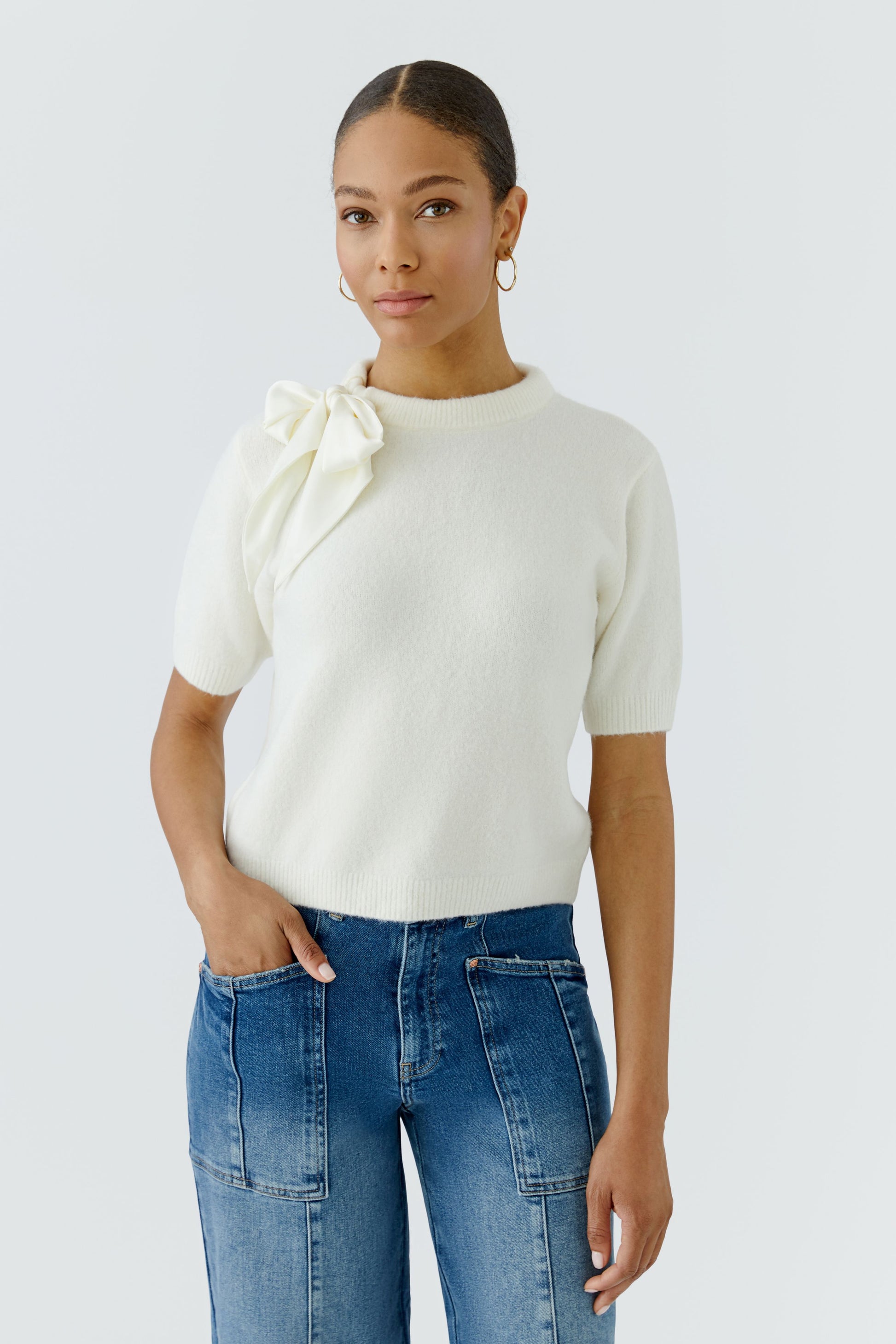 A woman modeling a cream short-sleeve knit top with a large bow detail on the shoulder, paired with blue jeans, against a light gray studio background.