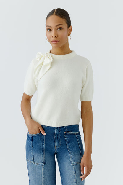 A woman modeling a cream short-sleeve knit top with a large bow detail on the shoulder, paired with blue jeans, against a light gray studio background.