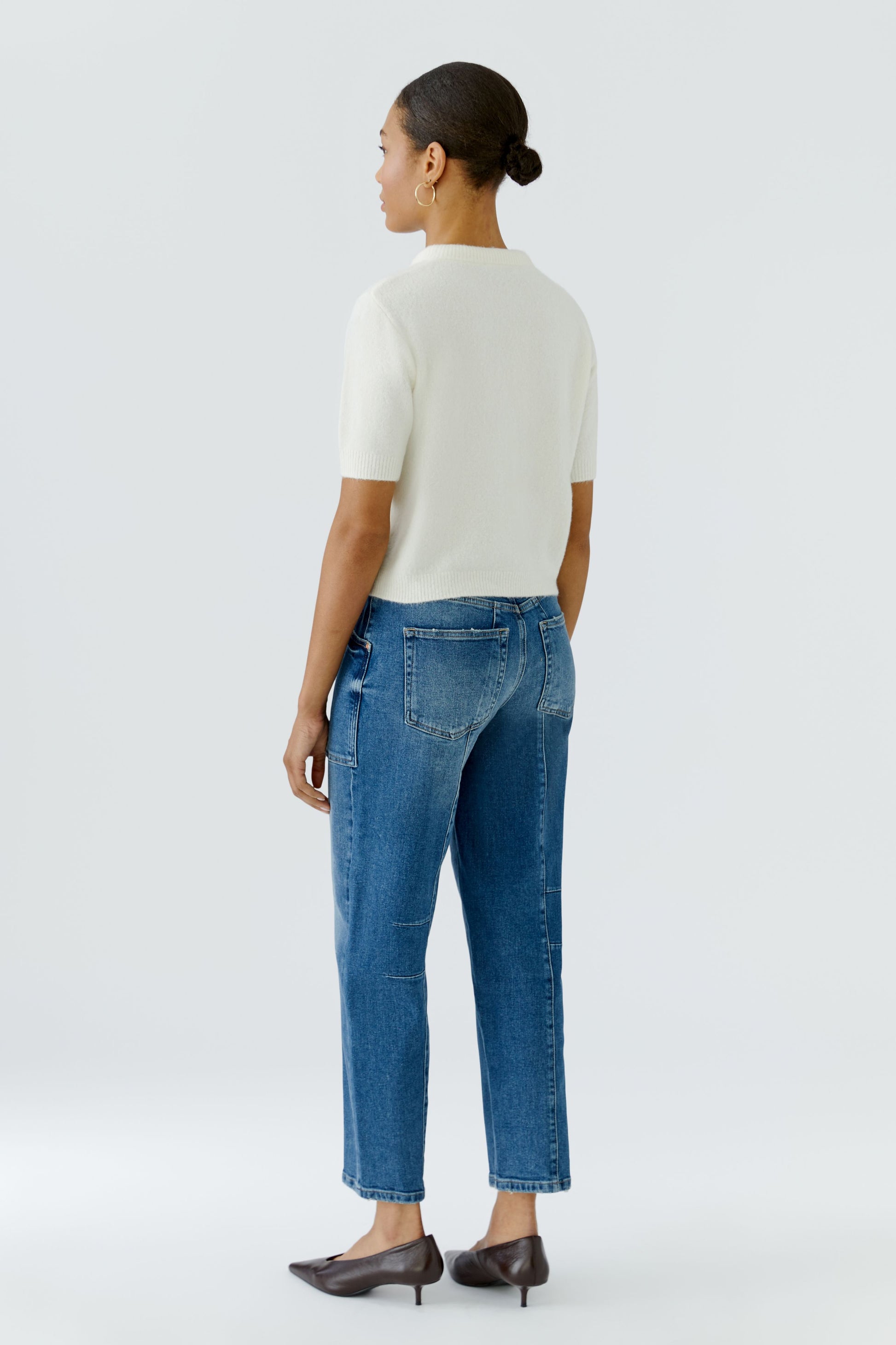 A woman shown from the back wearing a white short-sleeve top, blue cropped jeans, and dark brown pointed-toe heels against a light gray studio background.