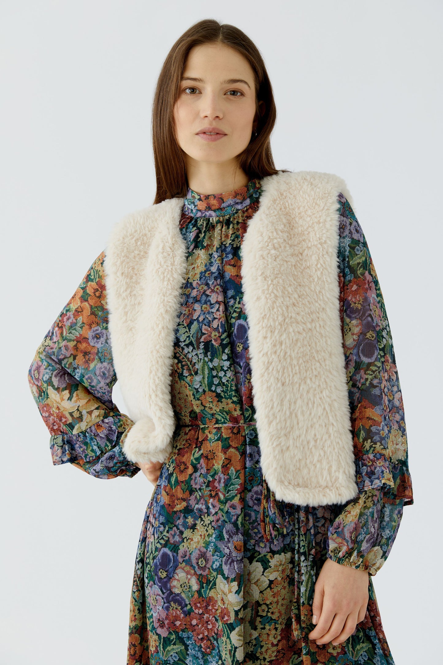 A woman wearing a cream-colored fuzzy faux-fur vest over a long-sleeved, multicolor floral print dress, photographed against a solid light gray background.