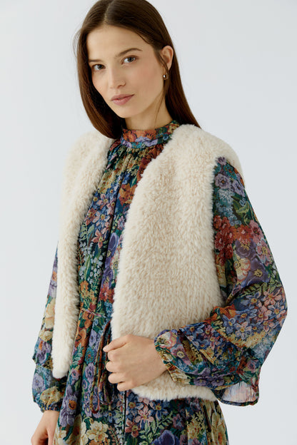 A woman wearing a cream faux-fur vest over a long-sleeve floral print blouse, photographed against a plain light gray studio background.