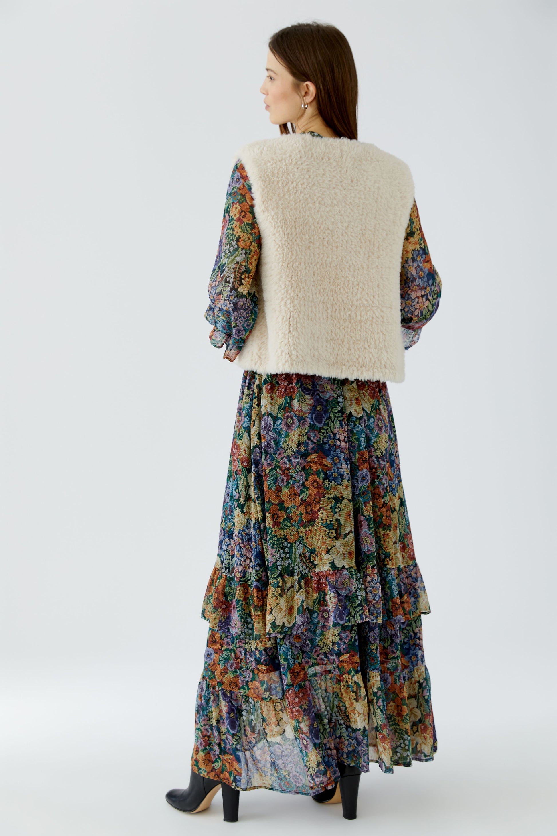 A woman shown from the back wearing a cream-colored textured vest over a long-sleeved, multicolor floral print dress with a tiered ruffle skirt, paired with black heels, against a light gray studio background.