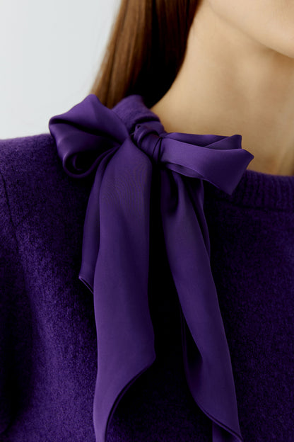 A close-up of a person wearing a deep purple knit sweater with a matching purple satin bow tied at the neckline.
