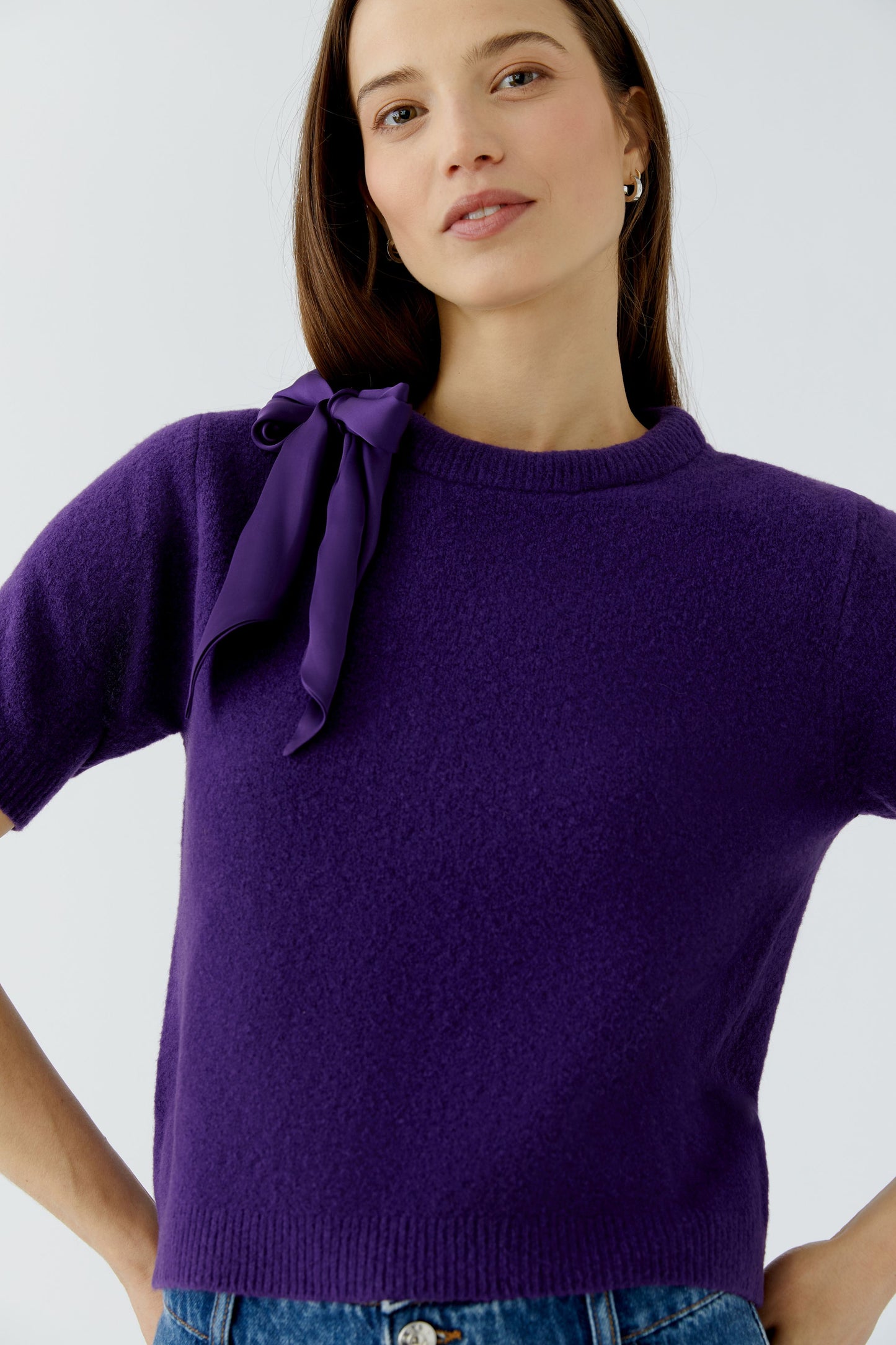 A woman wearing a short-sleeve purple knit sweater with a large bow detail on the left shoulder, paired with blue jeans against a light gray studio background.