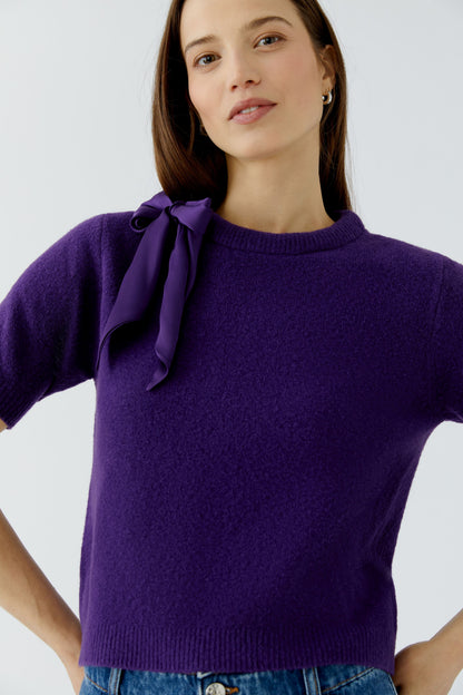 A woman wearing a short-sleeve purple knit sweater with a large bow detail on the left shoulder, paired with blue jeans against a light gray studio background.