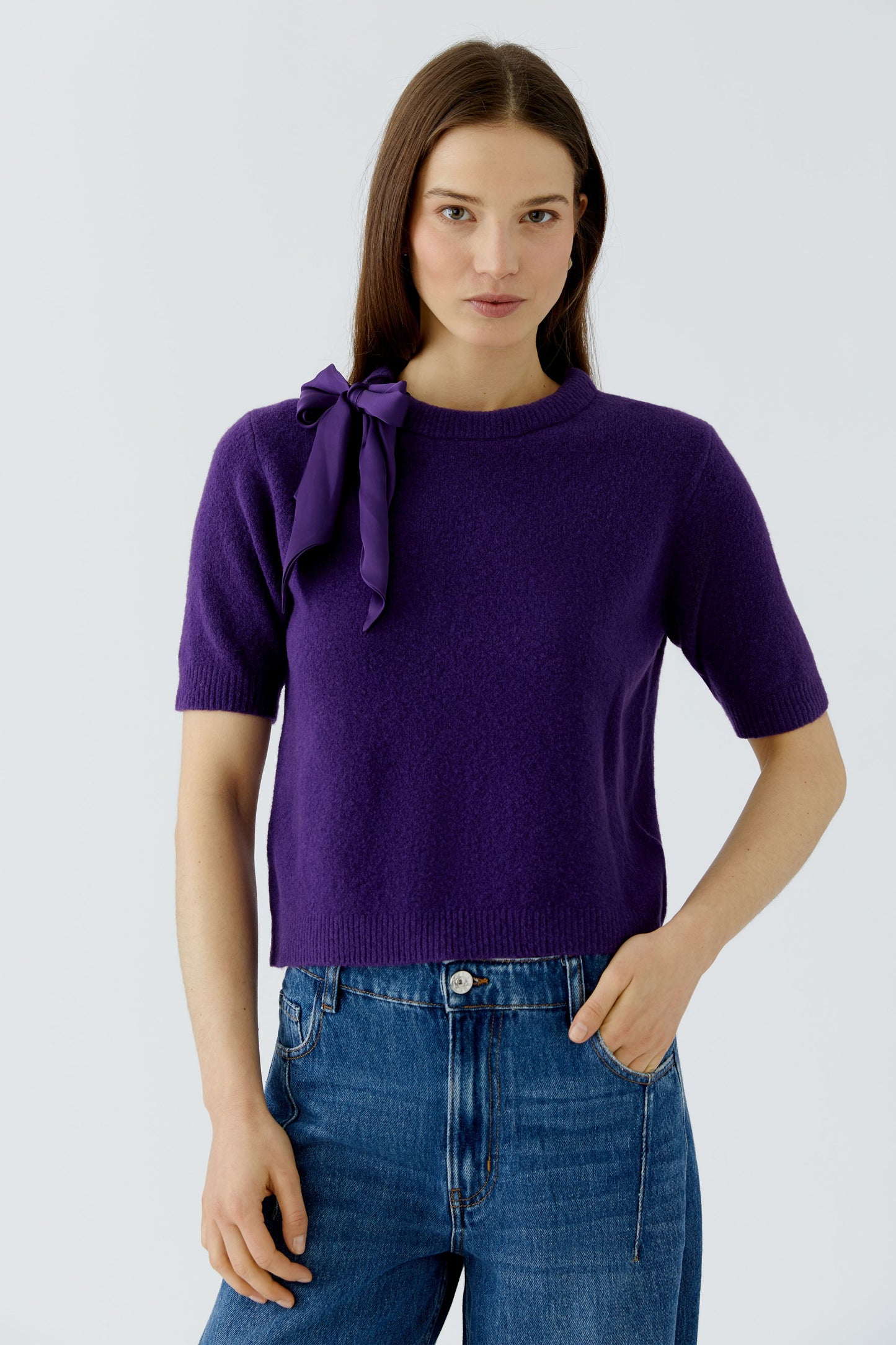 A woman modeling a short-sleeve purple knit top with a large bow detail on the left shoulder, paired with blue jeans, against a light gray studio background.