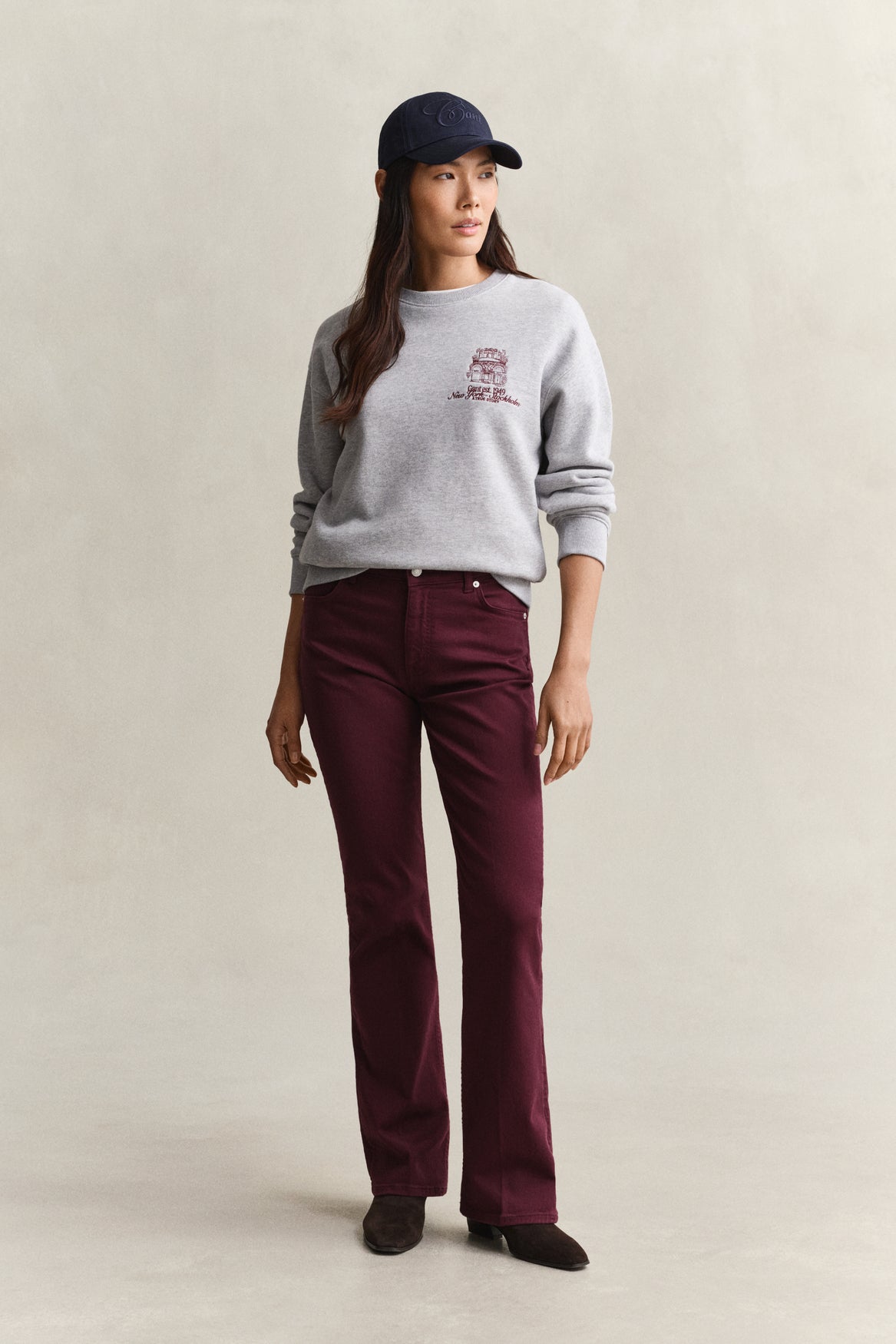 A woman stands against a plain light beige studio background wearing a light gray crewneck sweatshirt with a small maroon graphic on the left chest, dark burgundy pants, a navy baseball cap, and dark shoes.