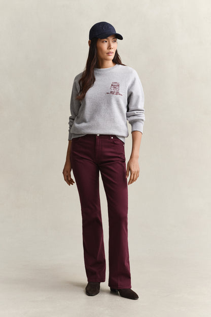 A woman stands against a plain light beige studio background wearing a light gray crewneck sweatshirt with a small maroon graphic on the left chest, dark burgundy pants, a navy baseball cap, and dark shoes.