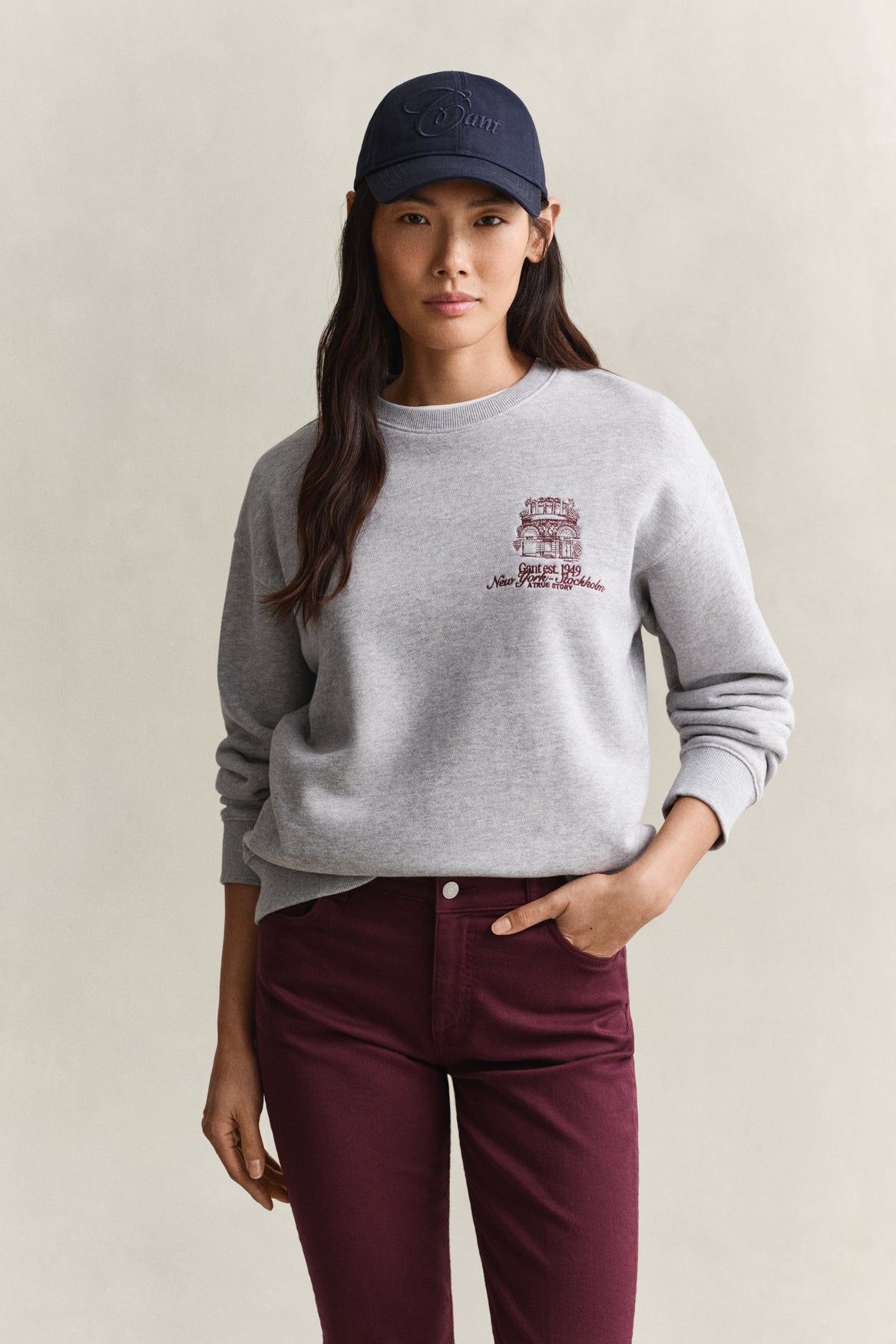 A woman modeling a light gray crewneck sweatshirt with a maroon graphic on the left chest, paired with dark burgundy pants and a navy baseball cap, against a neutral beige background.