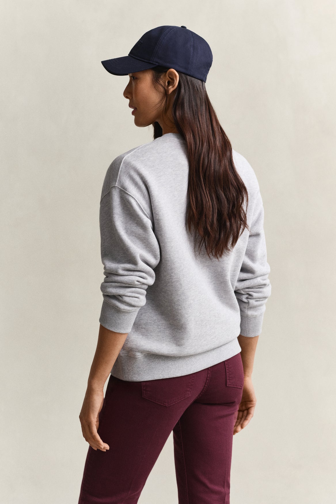 A woman shown from the back wearing a light gray crewneck sweatshirt, a navy blue baseball cap, and burgundy pants, set against a plain beige studio background.