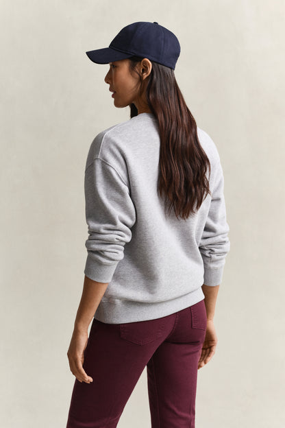 A woman shown from the back wearing a light gray crewneck sweatshirt, a navy blue baseball cap, and burgundy pants, set against a plain beige studio background.