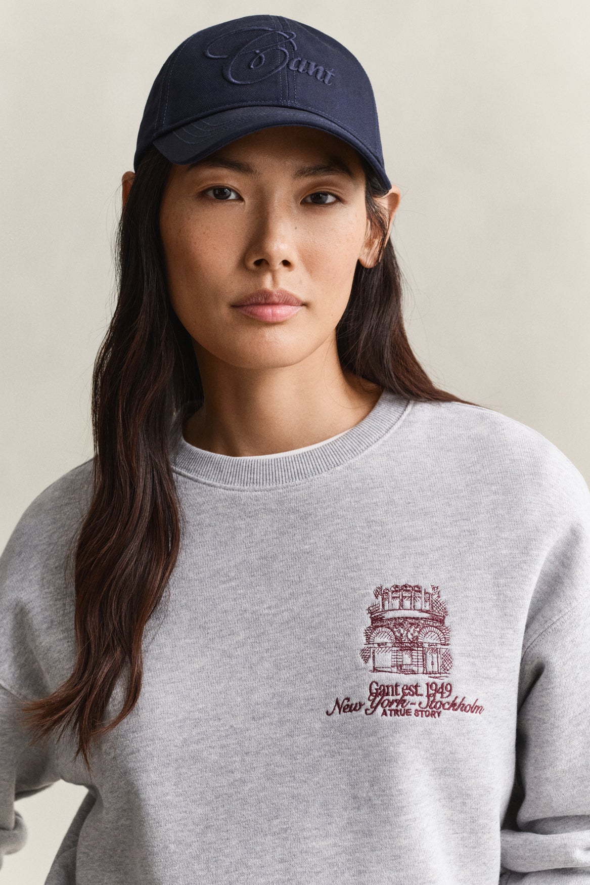 A woman wearing a heather gray crewneck sweatshirt with a burgundy New York & Company architectural graphic on the left chest, paired with a navy blue embroidered cap.