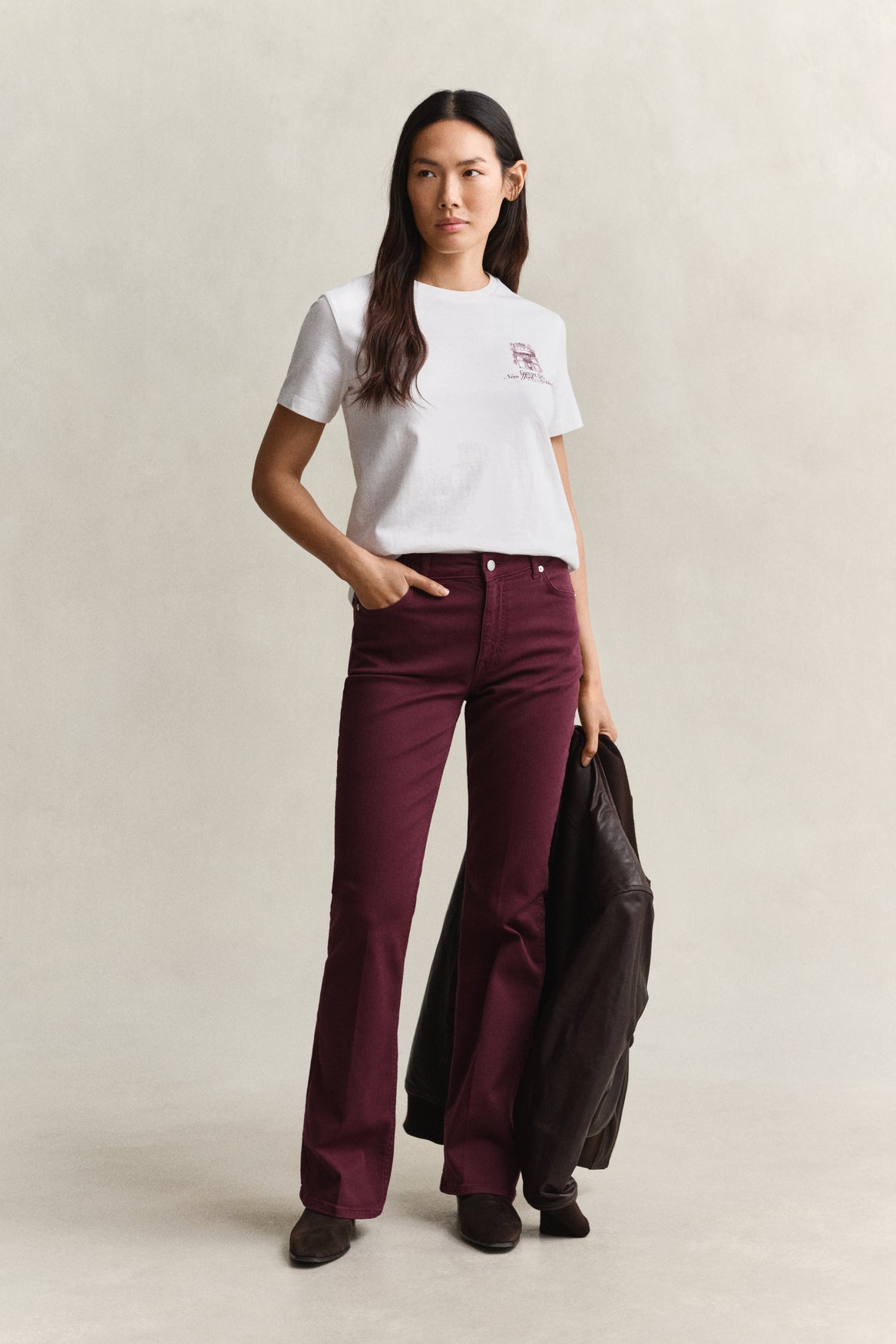 A woman stands against a plain light beige studio background wearing a white short-sleeve T-shirt with a small chest graphic, dark burgundy flare pants, and holding a black jacket. No visible brand text is legible.