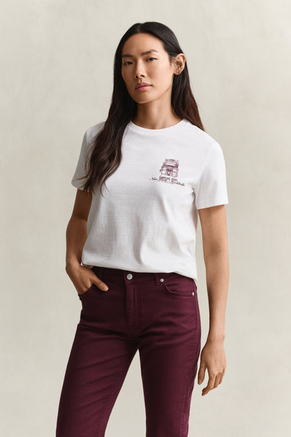 A woman modeling a white short-sleeve crewneck t-shirt with a small maroon graphic on the left chest, paired with dark burgundy pants, against a neutral beige background.