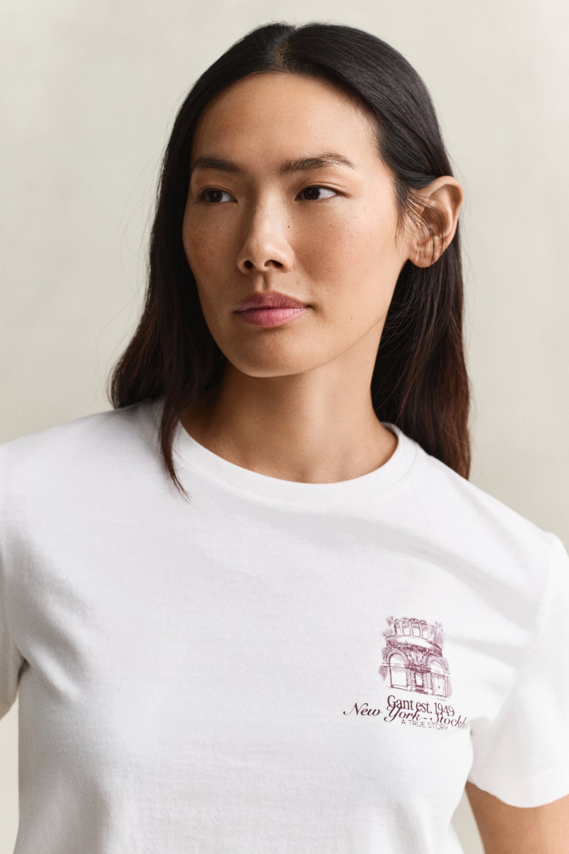 A woman wearing a white crew-neck t-shirt with a small maroon graphic of the New York City Hall and the text 'Garden City, New York' on the left chest, set against a plain light beige background.