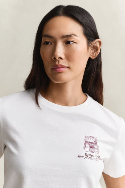 A woman wearing a white crew-neck t-shirt with a small maroon graphic of the New York City Hall and the text 'Garden City, New York' on the left chest, set against a plain light beige background.