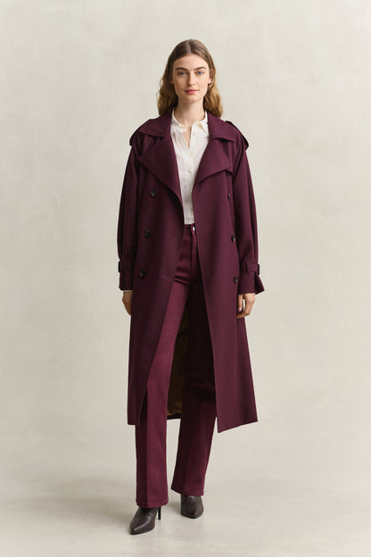 A woman modeling a long, double-breasted trench coat in deep burgundy, worn over a white collared shirt and matching burgundy trousers, against a plain light beige studio background.
