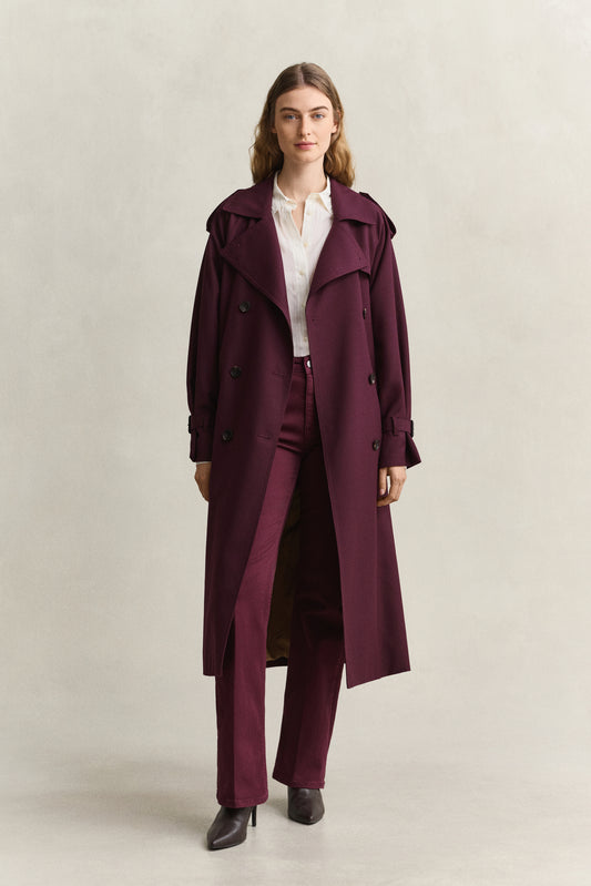 A woman modeling a long, double-breasted trench coat in deep burgundy, worn over a white collared shirt and matching burgundy trousers, against a plain light beige studio background.