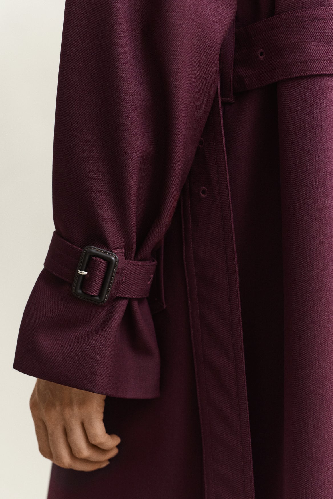 Close-up of a burgundy trench coat with a matching belt and buckle detail on the sleeve, worn by a model.