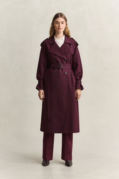 A woman stands against a plain light beige studio background wearing a long burgundy trench coat with a matching belt and wide-leg trousers, layered over a white collared shirt.