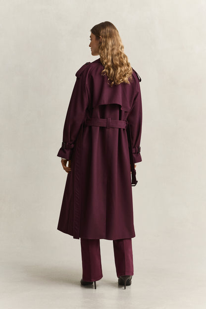 A woman shown from the back wearing a long burgundy trench coat with a matching belt, matching trousers, and black heels against a plain light gray studio background.
