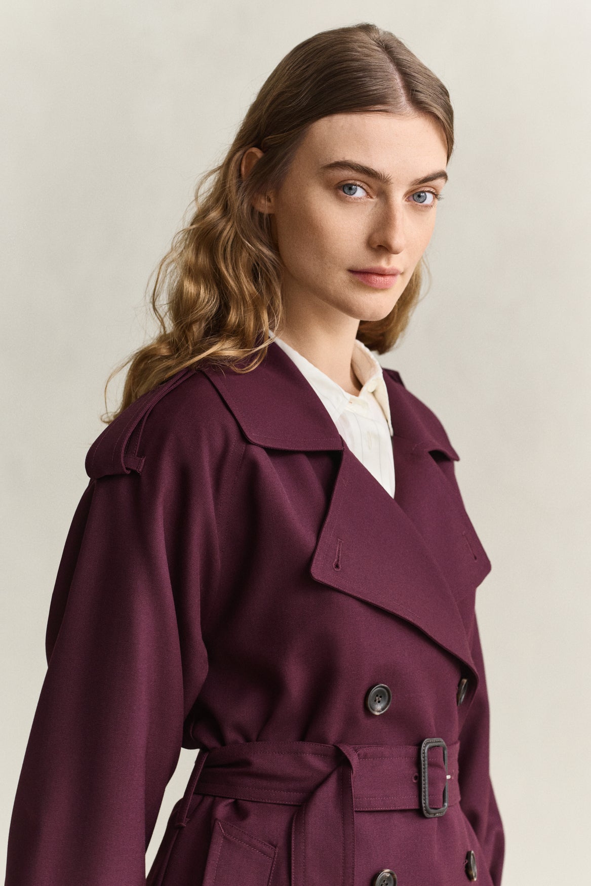A woman wearing a double-breasted belted trench coat in deep burgundy over a white collared shirt, photographed against a plain light beige studio background.